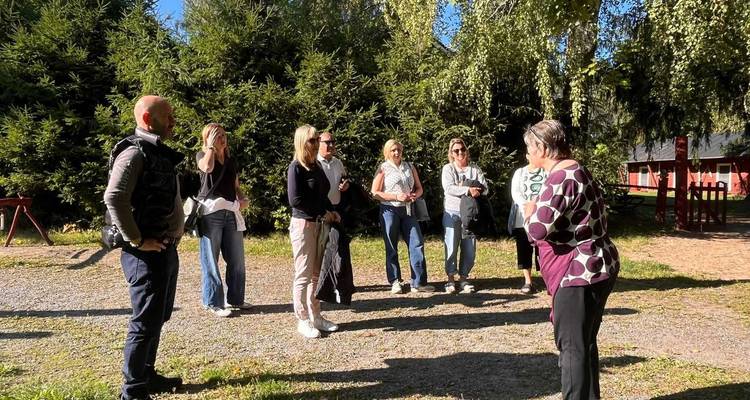 A guide speaks to a small group standing on grassy ground surrounded by evergreen trees in bright light.