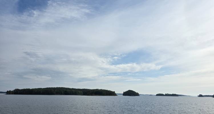 Calm lake waters stretch to the horizon dotted with low, forested islands beneath a lightly clouded sky.