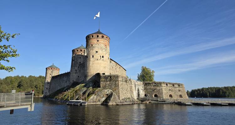 Stone Olavinlinna Castle rises from water atop a rocky islet under a clear blue sky.