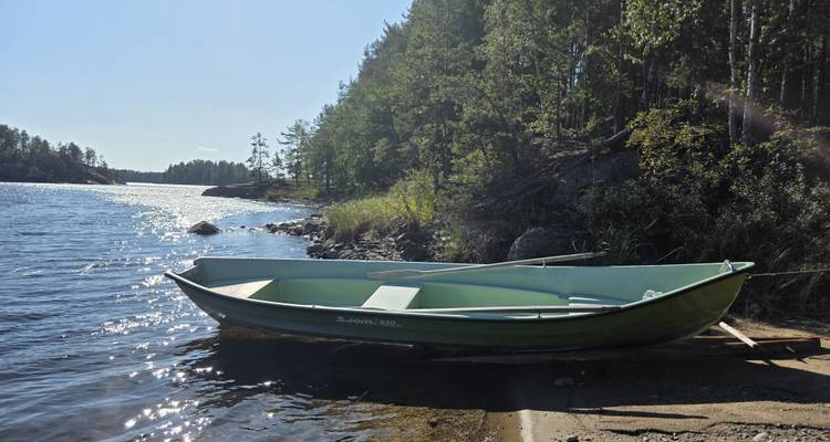 A green rowboat rests on a sandy shore beside a sparkling forest lake under bright sun.