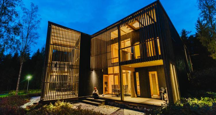 Contemporary wooden lodge glowing warmly at dusk with one person sitting on the steps.