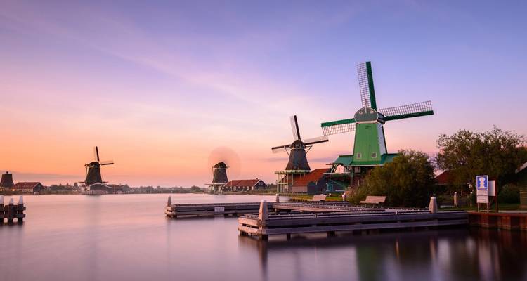 Iconic Dutch windmills line a tranquil canal under a pastel sunrise sky at Zaanse Schans