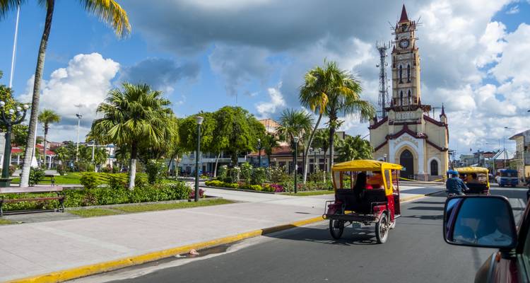 Small moto-taxis drive past a cathedral and landscaped plaza under patchy clouds.
