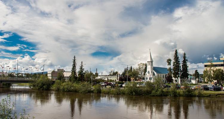 A small riverside cityscape with a white-steepled church and parked cars reflected in calm brown water beneath a partly cloudy sky.
