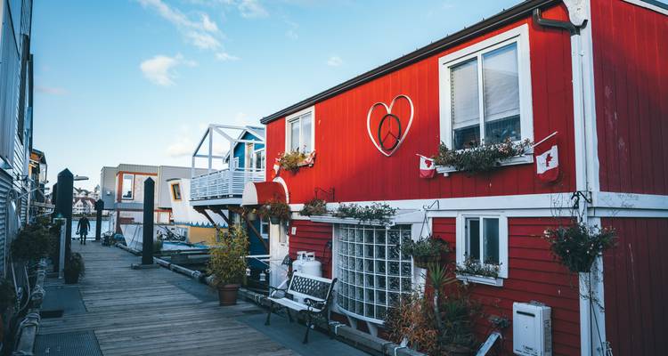 Colorful floating homes line a wooden boardwalk under bright morning light, with Canadian flags decorating a red house.