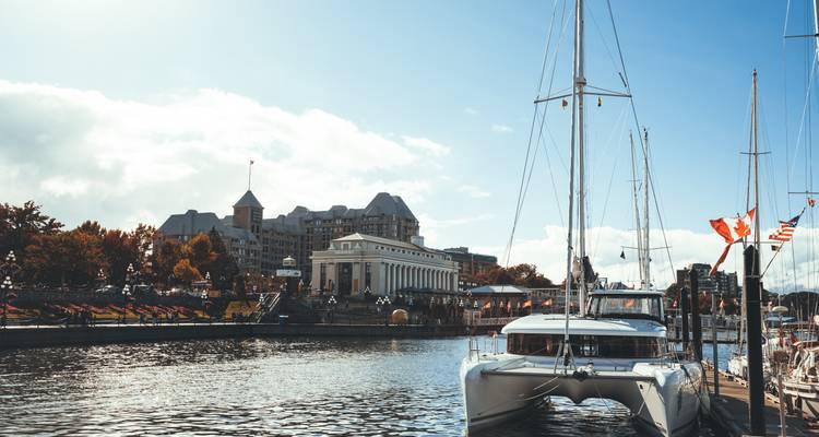 Sailboats are docked beside waterfront promenades and heritage buildings under a bright blue sky with Canadian flags fluttering.
