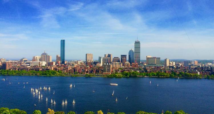 Boston skyline rises over the Charles River where scores of small sailboats glide across the blue water.