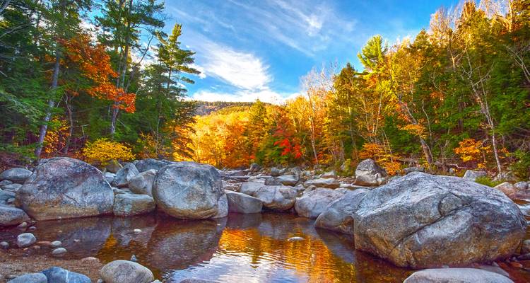 Clear mountain stream flows between large boulders amid a brightly colored autumn forest under blue sky.