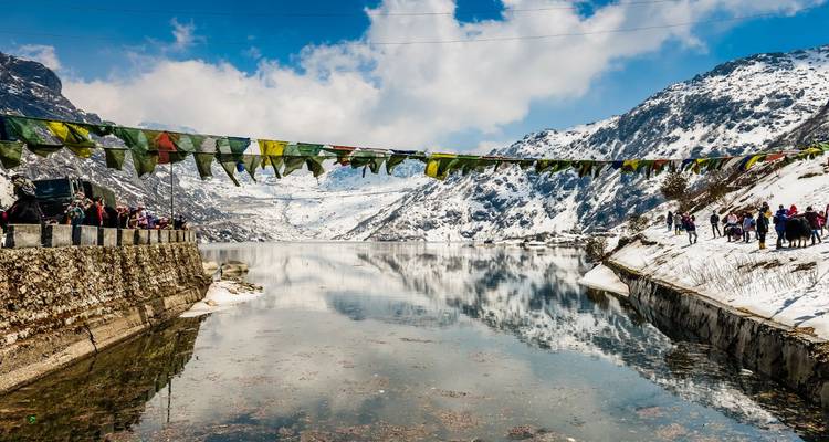 Clear alpine lake ringed by snow-covered mountains, mirrored reflections and a strand of prayer flags with visitors on the shore.