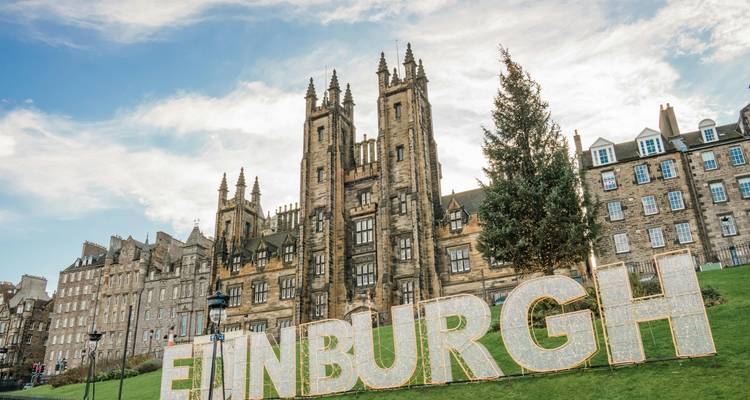 Des lettres géantes illuminées formant le mot ÉDIMBOURG sont placées devant un imposant bâtiment de style Gothic Revival sous un ciel dégagé.