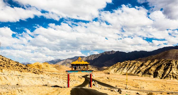 Lonely road curving through barren mountains toward a brightly painted gateway under scattered clouds.