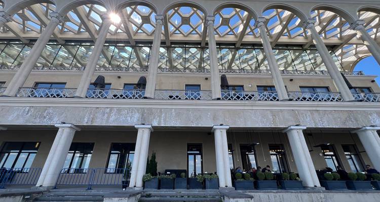 Grande terrasse à colonnades avec arches ornementées et toit de verre d'un bâtiment de vinerie contre un ciel lumineux.