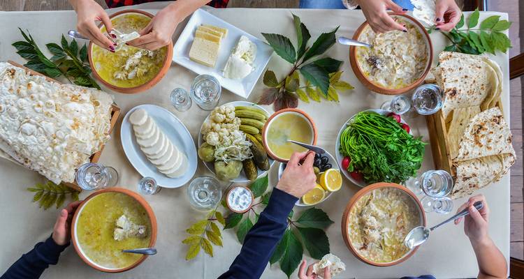 Vue de dessus d'une table présentant des plats arméniens, des herbes fraîches, du pain lavash et des convives partageant un repas.