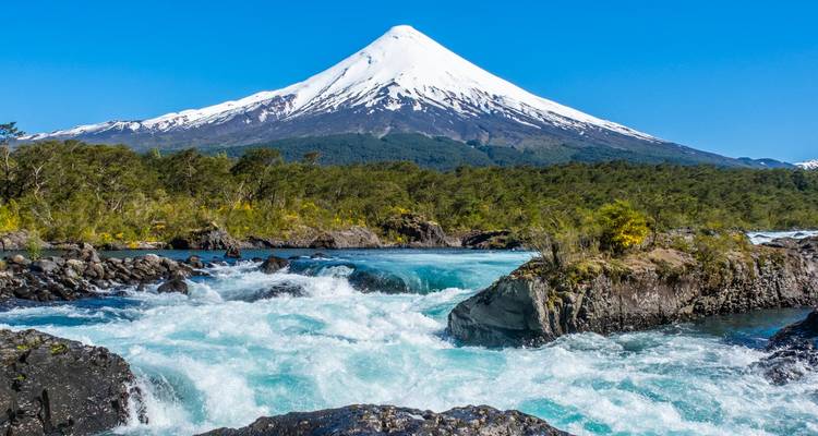 Raging turquoise river with snowy volcano towering above dense green forest under a clear sky.