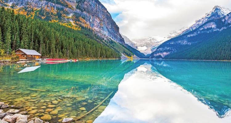 Crystal-clear turquoise water reflects snow-capped Rocky Mountains and dense evergreen forest beside a small wooden boathouse.