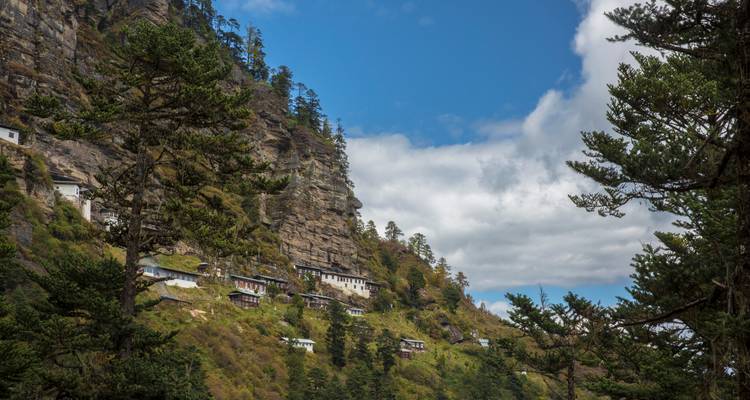 Clustered hermitages perch on a forested cliff beneath a bright blue sky and drifting clouds.