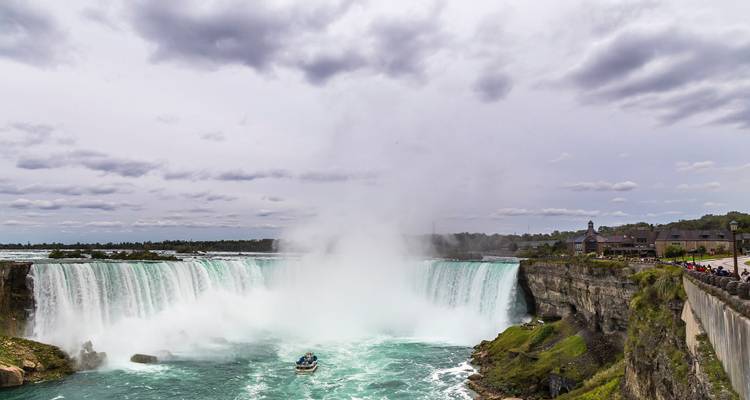 Powerful Niagara Falls plunges into turquoise rapids while mist rises high into a cloudy sky.