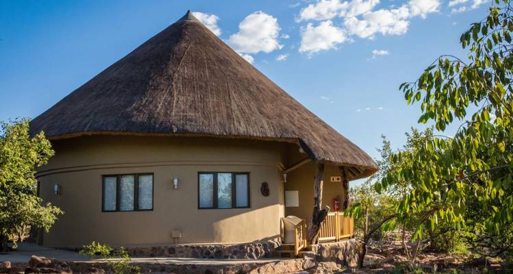Round thatched-roof chalet lodge nestled among bushveld under clear blue sky.