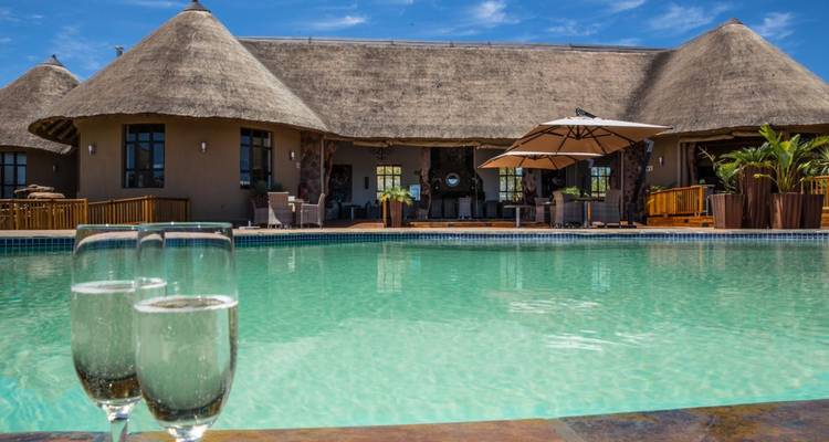 Sparkling glasses of champagne sit on ledge beside turquoise swimming pool overlooking luxury thatched lodge.