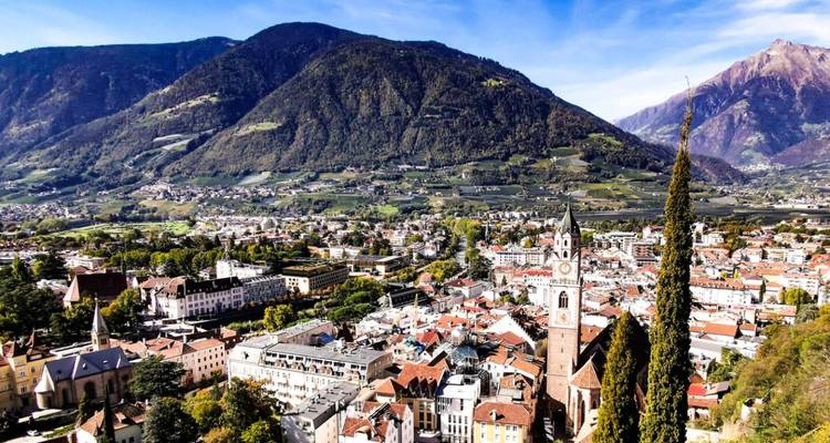 Uitgebreid luchtfoto van Merano's terracotta daken tegen beboste bergen onder een heldere hemel.