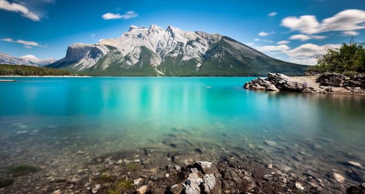 Long-exposure image of a glassy turquoise lake edged by rocky shoreline with dramatic Rocky Mountain peaks reflected in the water.