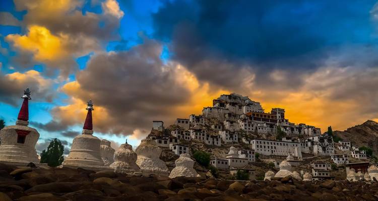 Whitewashed hilltop monastery and stupas under dramatic sunset clouds in Ladakh