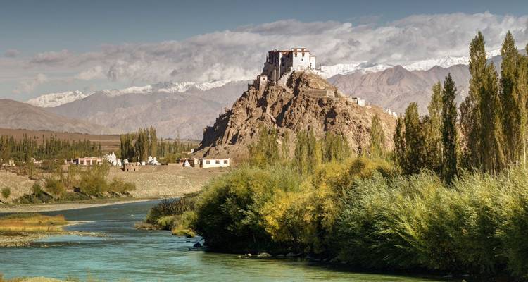Castle-like monastery perched on a rocky hill above a winding river with snow peaks behind