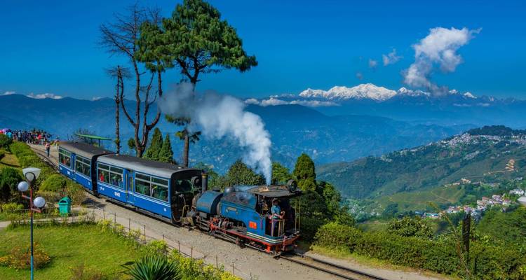 A blue steam train chugs along a hillside track with snow-capped Himalayan peaks in the distance.