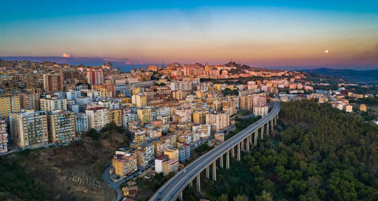 Aerial panoramic of a hillside Sicilian city with a long viaduct at sunset and rising moon.