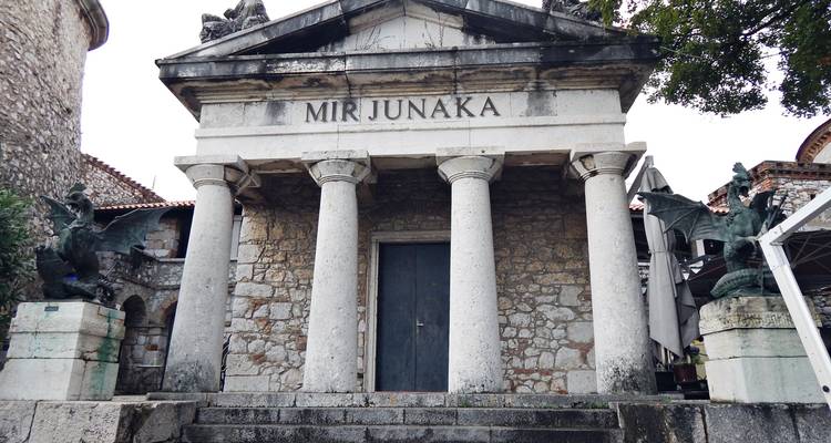 Stenen mausoleum met vier klassieke zuilen en het opschrift "MIR JUNAKA" geflankeerd door drakenbeelden.