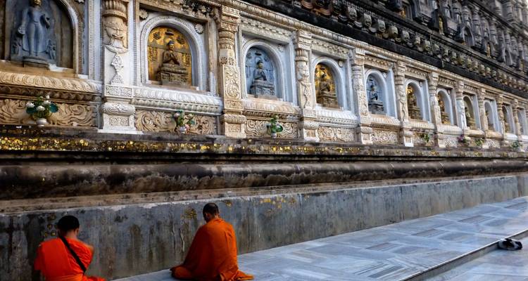 Two monks in saffron robes praying along an intricately carved stone relief wall.