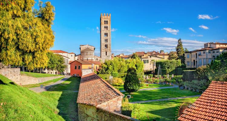 Lush gardens and medieval tower rising above terracotta rooftops in a walled Italian city.