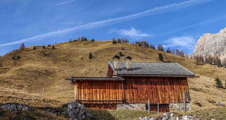 Cabaña alpina de madera situada contra una ladera de hierba seca con un cielo azul claro surcado por una sola estela de condensación.