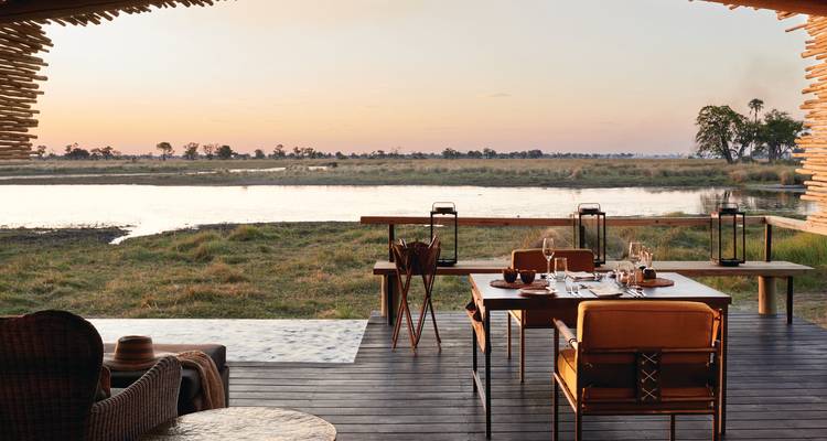Stijlvol openlucht lodge terras gedekt voor diner met uitzicht op een rustige rivier bij zonsondergang.