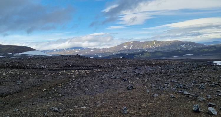 Plaine volcanique caillouteuse s'étendant vers des crêtes lointaines sous un mélange de nuages et de soleil.