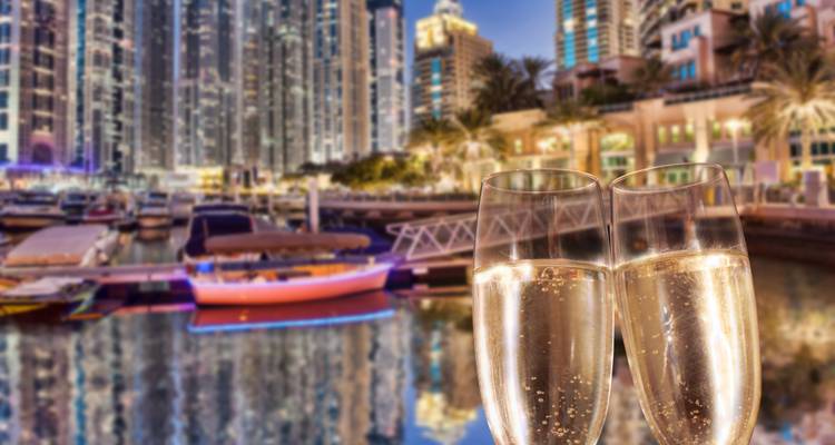 Nahaufnahme von zwei Champagnerflöten mit Sekt vor einer unscharfen nächtlichen Skyline und einem Yachthafen.