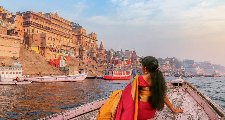 Frau in Sari sitzt in einem Boot und bewundert die antike Flussufferarchitektur von Varanasi bei Tagesanbruch.
