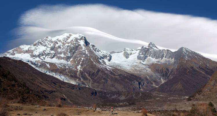 Schneebedeckte Himalaya-Kette, die sich über ein breites Tal unter einer dramatischen Wolkenkappe erhebt.