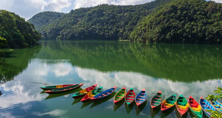 Reihe von leuchtend bemalten Holzbooten, die auf dem smaragdgrünen Wasser des Phewa Lake ankern und von grünen Hügeln umgeben sind.