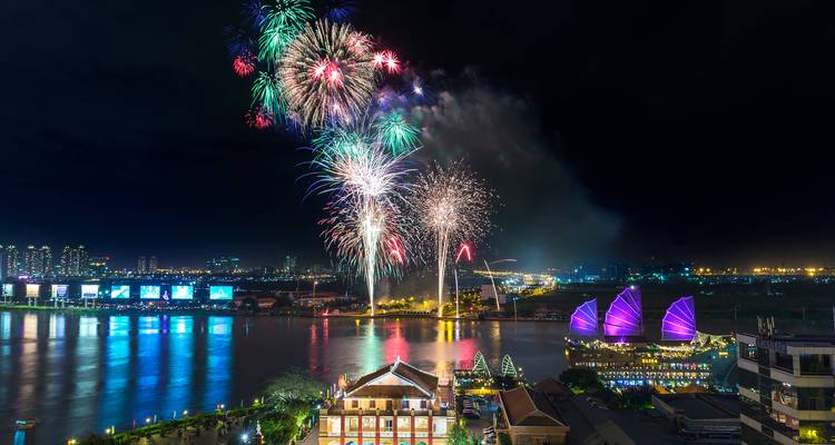 Colourful fireworks burst above a riverside skyline featuring illuminated purple sails.