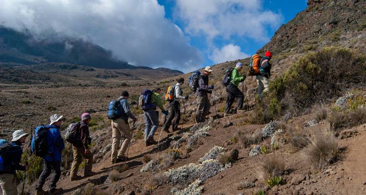 Línea de excursionistas ascendiendo por una ladera volcánica árida del Kilimanjaro bajo nubes dramáticas.