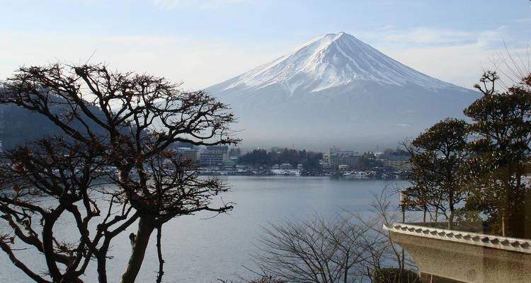 Der schneebedeckte Berg Fuji spiegelt sich in einem ruhigen See wider, umrahmt von Winterbäumen und einer fernen Stadt.