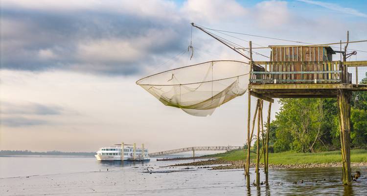 Traditional wooden fishing hut with large net over a calm estuary as a river cruise ship approaches.