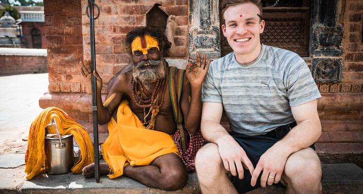 A tourist sits beside a colourfully painted sadhu who gestures peace signs at a temple courtyard.
