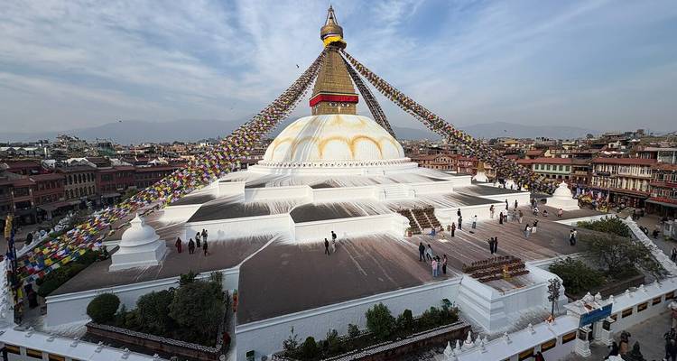 Wide panoramic view of Boudhanath Stupa with hundreds of prayer flags and visitors walking below.