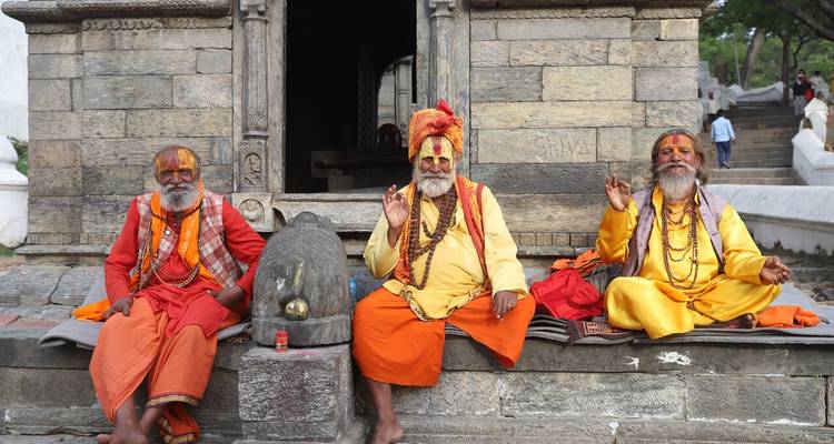 Three saffron-clad sadhus sit in front of a stone shrine, giving blessing gestures.