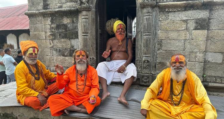 Group of colourfully painted Hindu ascetics sit outside a stone shrine, one elderly man behind them.