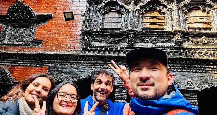 Four friends smile and flash peace signs beneath ornate wood-carved windows in Kathmandu.