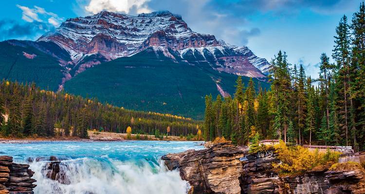 Powerful turquoise Athabasca Falls crashes in the foreground with snow-dusted Mount Kerkeslin rising above dense evergreen forest in Jasper National Park.