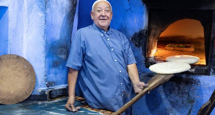Un panadero local metiendo barras de pan redondas en un horno tradicional de leña en una panadería pintada de azul.